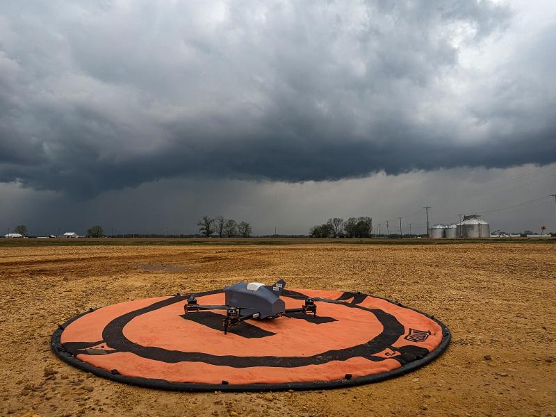 A gray uncrewed aircraft sits on a orange launch pad in an open field. A storm with dark clouds forms in the distance