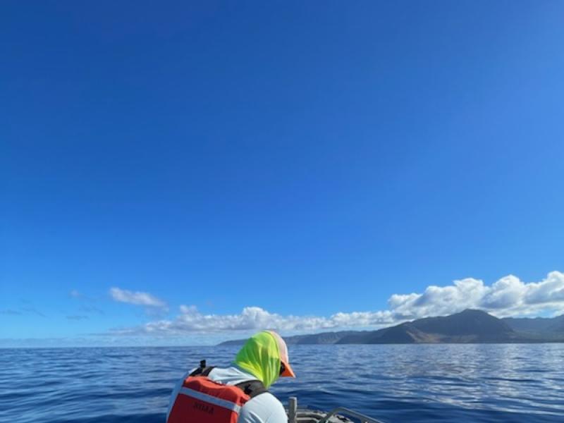 Two people hold on to yellow underwater gliders in the front of a small boat as they approach an island.