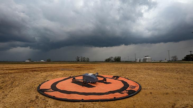 A gray uncrewed aircraft sits on a orange launch pad in an open field. A storm with dark clouds forms in the distance
