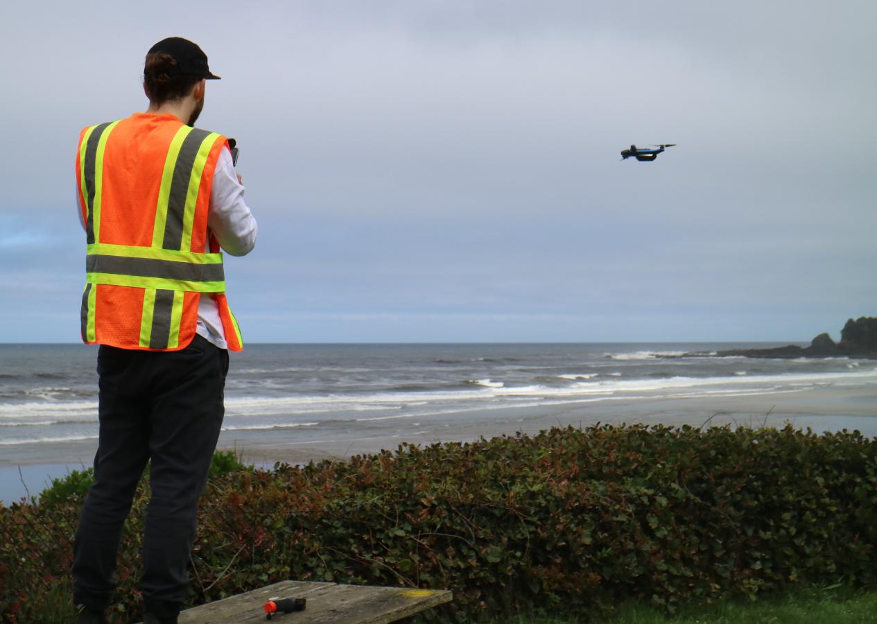 A man in an orange and yellow safety vest stands on a beach with his back to the camera. He is piloting a small drone that is flying in the distance over the sand.