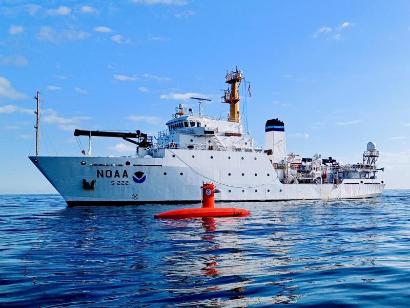 A white NOAA ship with an orange uncrewed surface vehicle on the water in the foreground