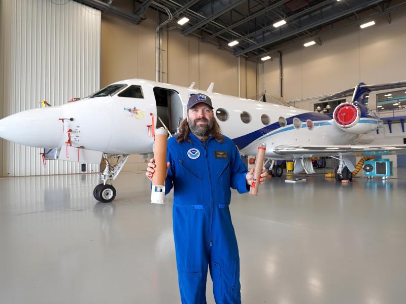 An engineer in a flight suit holds up two weather probes called dropsondes