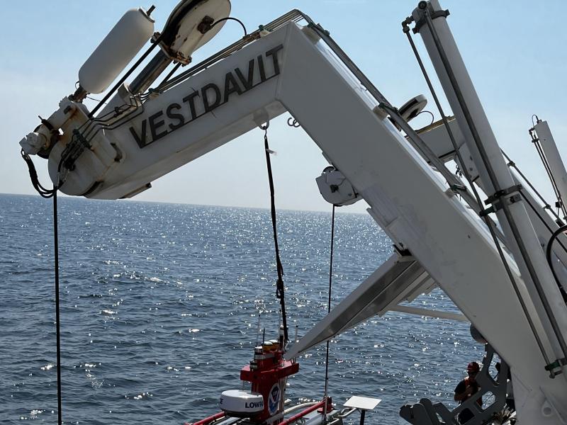 A gray davit arm extends over the edge of a ship. The DriX deployment gondola with a red DriX are being lowered by the davit.