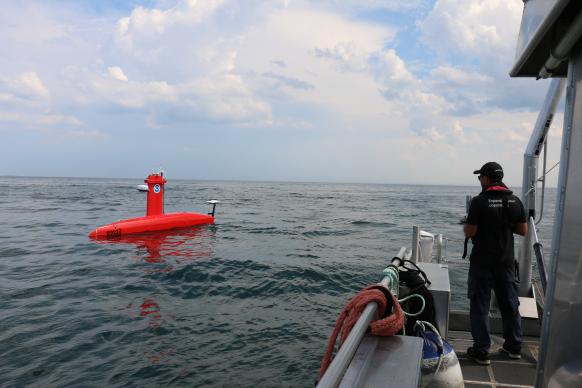 An operator stands on the side of a small boat as it pilots a red DriX uncrewed surface vehicle in the distance.