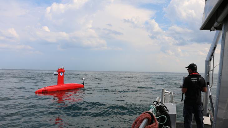 An operator stands on the side of a small boat as it pilots a red DriX uncrewed surface vehicle in the distance.