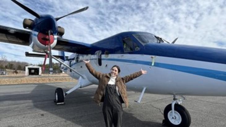 An OMAO Knauss Fellow stands in front of a NOAA Twin Otter plane, posing with a smile and outstretched arms.