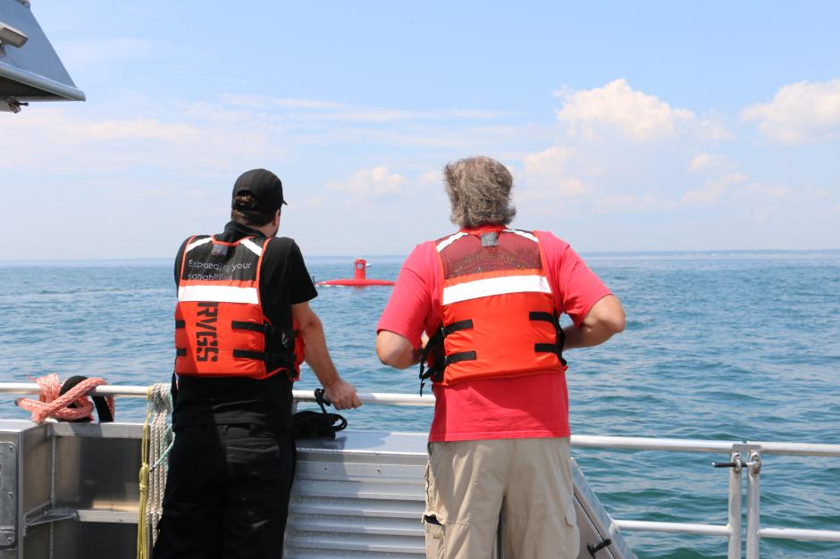 Two people in orange life jackets with their backs to the camera stare out at the ocean as a red uncrewed surface vehicle drives by.